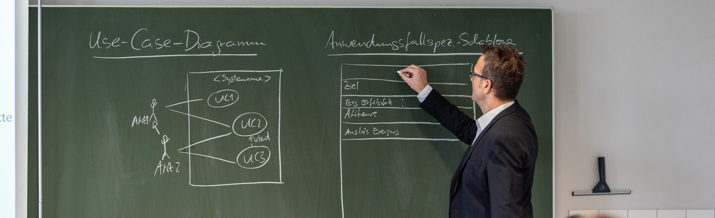 Photo of a professor writing something on the blackboard in the computer room. In front of him some students at computers.