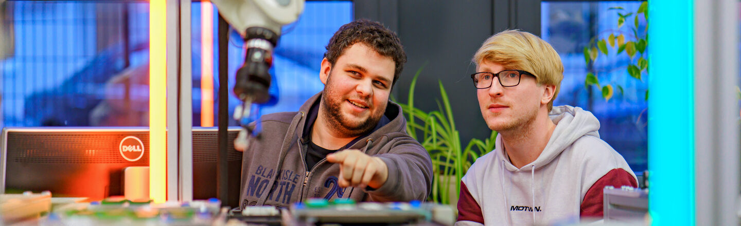 Foto von zwei Studierenden, die hinterm Transportband sitzen. Der linke Studierende zeigt zum Roboterarm vor ihnen.