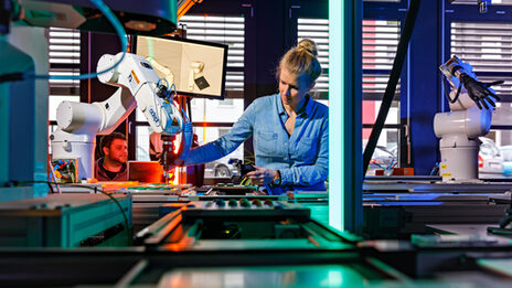 Photo of a conveyor belt with robot arms behind which an employee stands who reaches for a robot arm with her right hand. __ Behind a conveyor belt with robot arms stands an employee who reaches for a robot arm with her right hand.