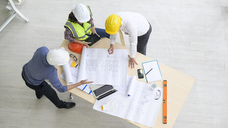 3 Ingenieur*innen in einer Besprechung an einem Tisch mit Plänen und Endgeräten