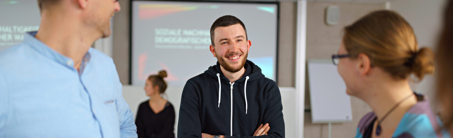 Photo of three people standing together in a seminar room and smiling at each other.