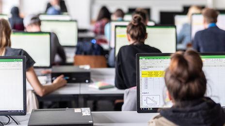 View from behind into the computer room with students, some Excel spreadsheets are visible on the monitors.