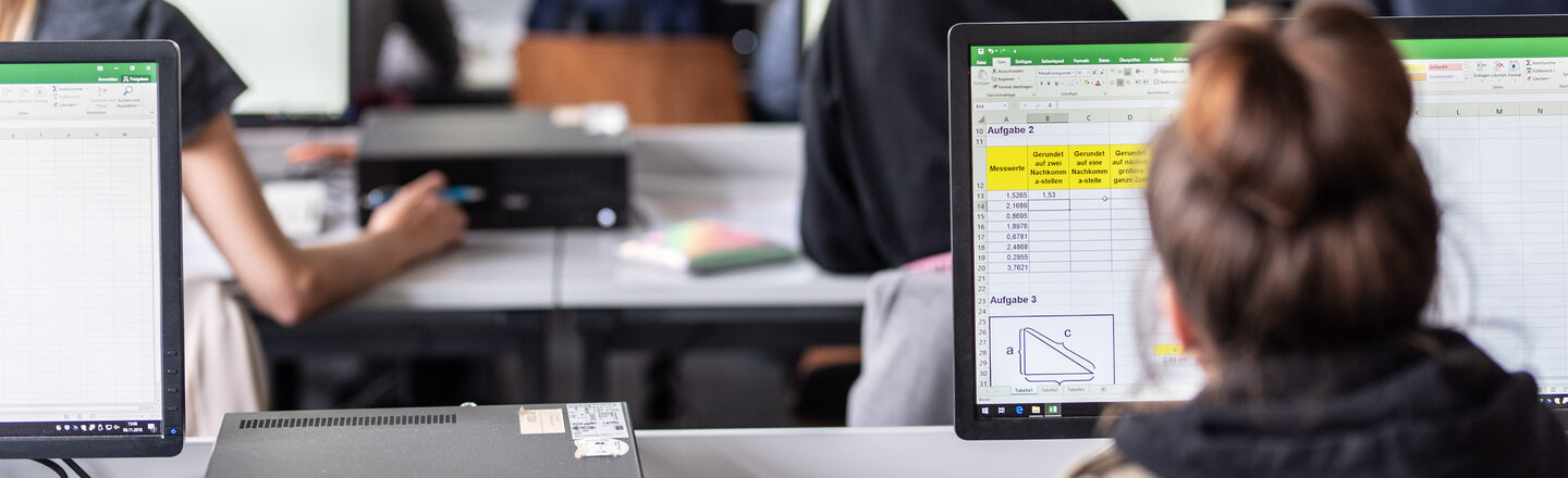 View from behind into the computer room with students, some Excel spreadsheets are visible on the monitors.