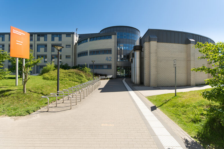 Photo of the building at Emil-Figge-Strasse 42 with a walkway with a guidance system for the blind. On the left a sign with the inscription "Fachhochschule Dortmund - Faculty of Computer Science".