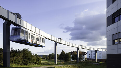 Foto von H-Bahn-Schienen mit vorbeifahrender H-Bahn. Rechts im Bild ein Gebäude im Anschnitt.