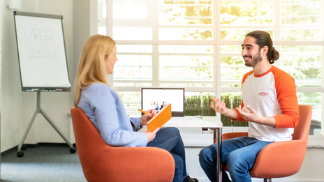 Photo of two people talking at a table. The employee smiles at the young man sitting opposite her and takes notes; she has her back to the camera. The young man beams and talks with his hands. A laptop can be seen on the table between the two of them and a flipchart can be seen blurred in the background on the left.