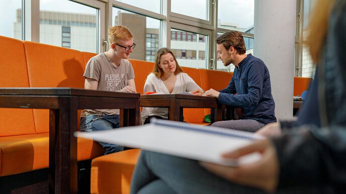 Foto einer Studentin und zweier Studenten auf orangefarbenen Sitzmöbeln. Sie betrachten gemeinsam Unterlagen auf dem Tisch. Rechts im Bild eine weitere Studentin im Anschnitt.__One female and two male students on orange chairs, they look together at documents on the table. On the right in the picture another student in the cut.