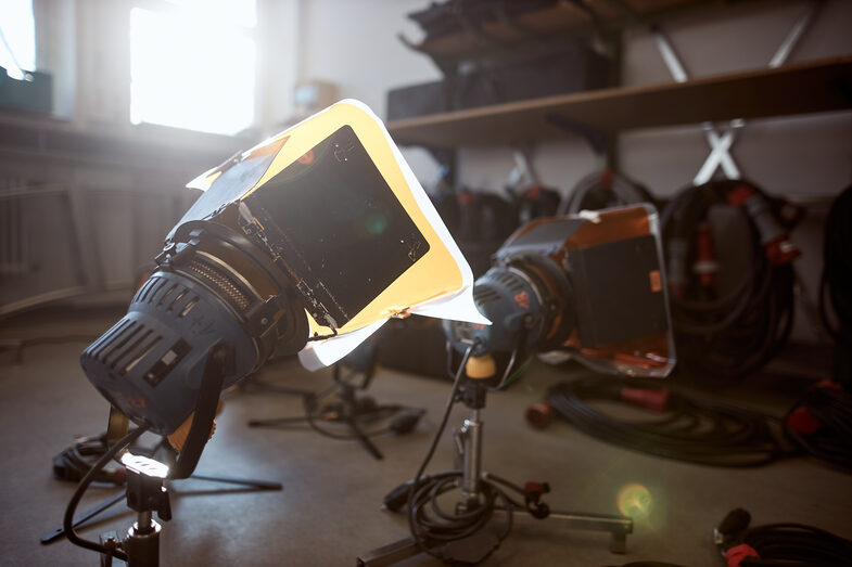 Foto einer Lampe mit orangefarbenen Diffusionsfilter, die auf dem Boden steht, dahinter steht eine weitere Leuchte. __ A lamp with an orange diffusion filter is on the floor, behind it is another lamp.