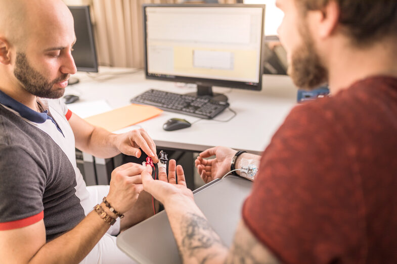 Foto zweier Männer am Schreibtisch. Der eine bringt bei dem anderen Elektroden an die Finger an, um seine Augenbewegung mittels Eyetracking zu messen