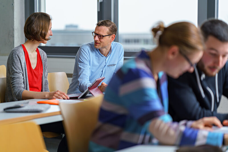 Photo of two people sitting next to each other in a seminar room and talking. Two other people in the foreground.