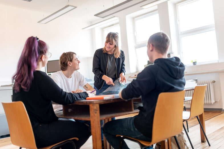 Foto von 4 Personen in einer Besprechung. Eine stehende Person zeigt einen Entwurf. Die 3 weiteren Personen sitzen an einem Tisch und halten Blickkontakt zur stehenden Person.