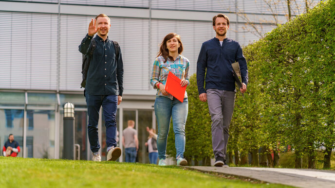 Foto von einer Studentin und zwei Studenten, die nebeneinander über den Campus gehen. Der eine Student winkt jemandem zu.__Three students walk side by side on a path, the student on the left raises his hand in greeting.