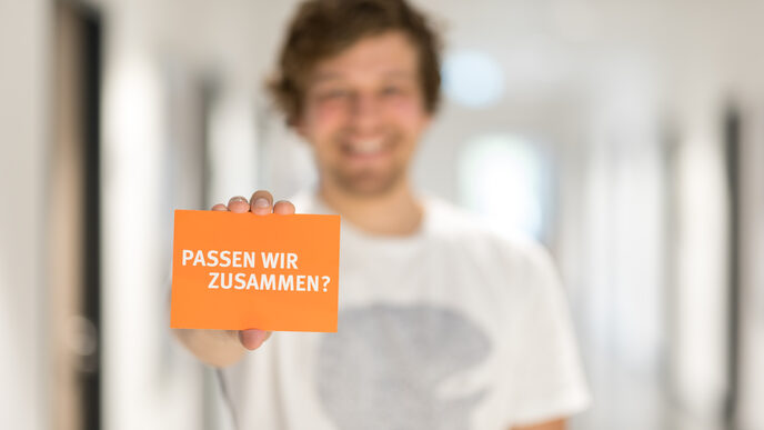 Foto eines Mannes, der eine Karte mit Schriftzug "Passen wir zusammen?" in die Kamera hält.__Man out of focus holding a card with the words "Do we fit together?" into the camera.