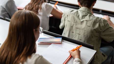 Foto einer jungen Frau, die sich Notizen im Hörsaal macht. Vor ihr sitzen drei weitere Personen. __ Young woman takes notes in the lecture hall, three other people are seated in front of her.