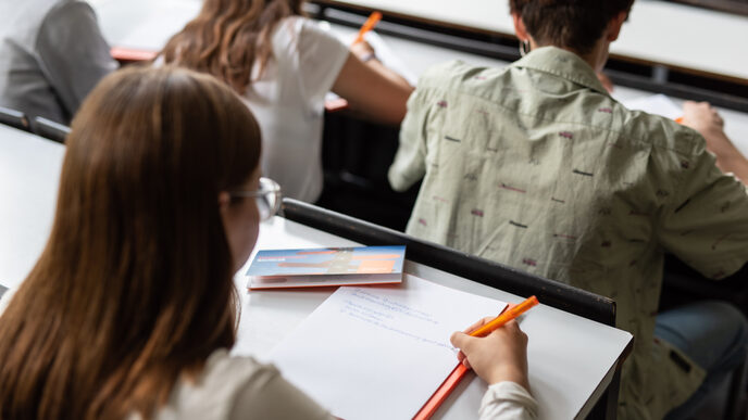 Foto einer jungen Frau, die sich Notizen im Hörsaal macht. Vor ihr sitzen drei weitere Personen. __ Young woman takes notes in the lecture hall, three other people are seated in front of her.