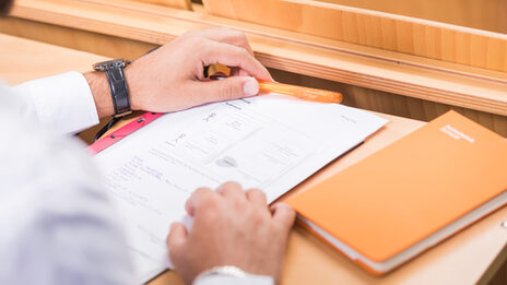 Foto über die Schulter einer Person an einem Tisch im Hörsaal. Auf dem Tisch liegen Zettel und ein Buch.