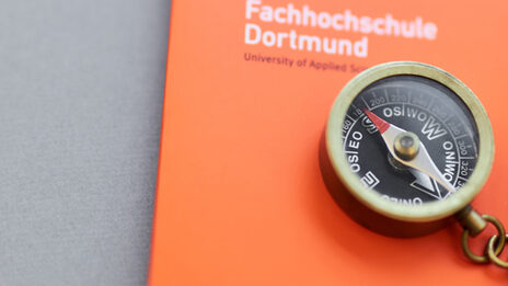 Photo of a small compass with a chain on it, lying with a pencil on an orange folder of the Fachhochschule Dortmund.