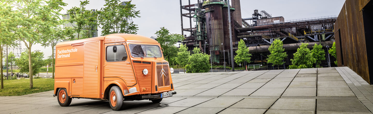 Foto von orangefarbenem Oldtimer-Kastenwagen mit FH-Logo vor Industriedenkmal.___Orange vintage car with FH logo in front of an industrial monument.