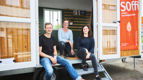 Man sieht 3 Personen, die in einem Bauwagen sitzen, welchen man über eine kleine Treppe betreten. An dem Bauwagen ist ein großes Plakat befestigt auf dem „Soffi mobiles Nachhaltigkeitsbüro“ steht.