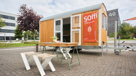Man sieht einen Bauwagen, mit einem Plakat auf dem „Soffi mobiles Nachhaltigkeitsbüro“ steht 
Vor dem Bauwagen sieht man einen Tisch auf dem ein paar Sachen liegen.