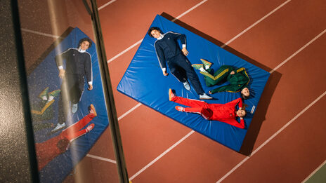 Das Foto zeigt drei Personen in einem Trainingsanzug, die auf einer Sportmatte liegen.