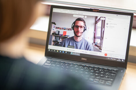 Foto über die Schulter einer Frau am Laptop, darauf läuft eine Videokonferenz mit einem Mann mit Headset.