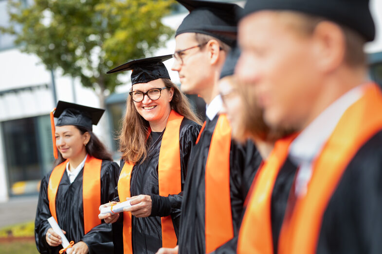 Foto von fünf Promovierenden, die in einer Reihe stehen und Talare und Doktorhüte tragen. Eine Promovierende lacht in die Kamera.