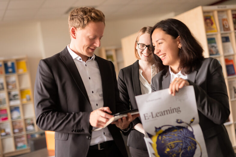 Foto von drei Promovierenden, die nebeneinander in der Bibliothek stehen. Eine junge Frau hält eine Zeitung mit der Aufschrift "E-Learning". Der Mann hält ein Smartphone, auf das er schaut.