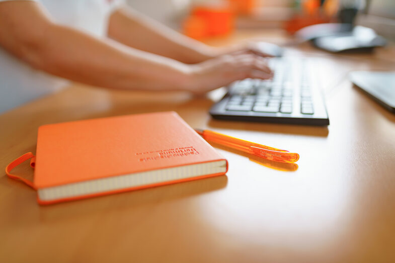 Photo of an FH notebook and FH ballpoint pen lying on a desk. A woman is typing on the keyboard in the background. __ FH notebook and FH ballpoint pen are on the desk, a woman is typing on the keyboard in the background.